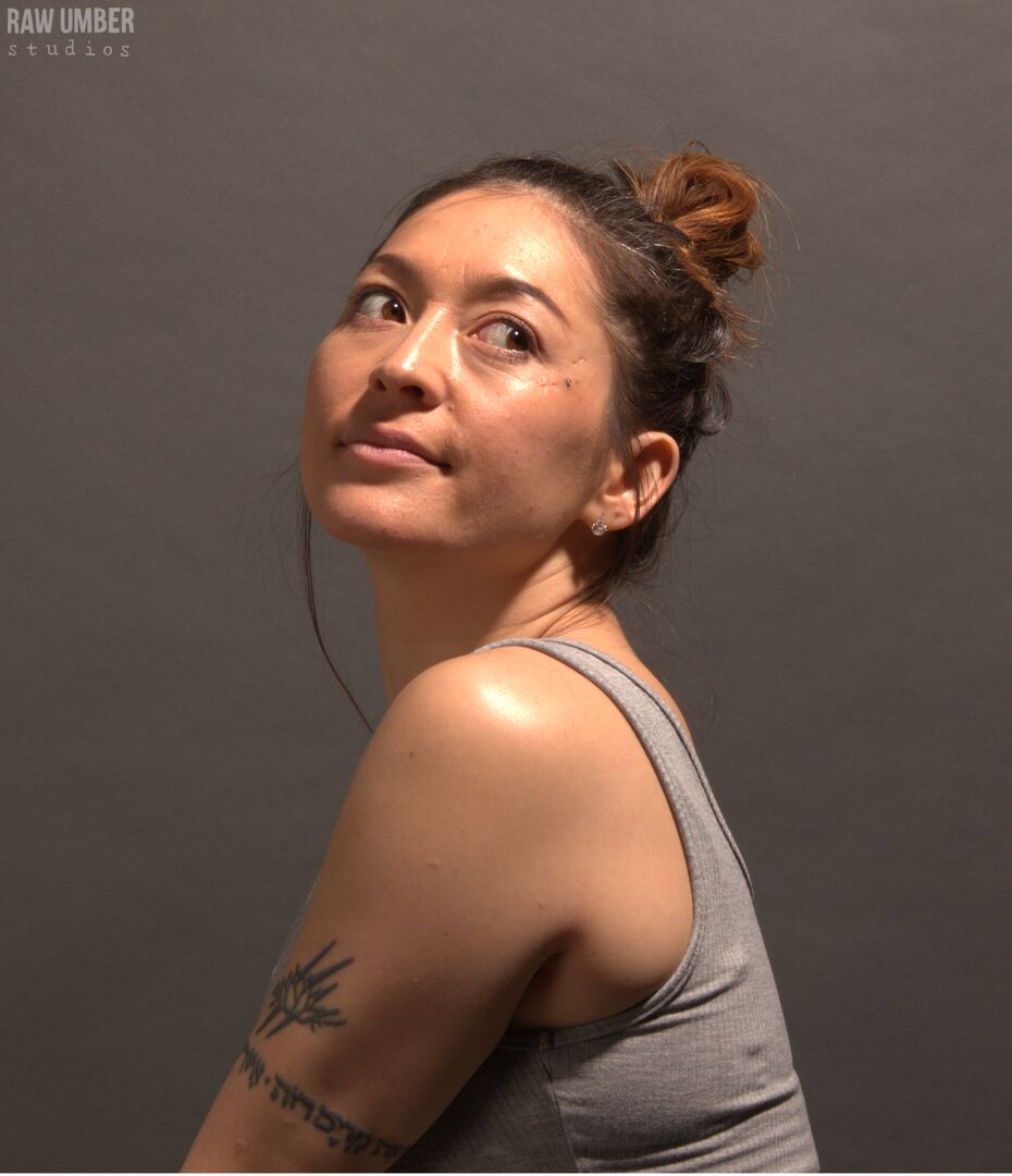 A woman with brown hair tied in a top knot and her body facing left looking over her left shoulder with a slight smirk