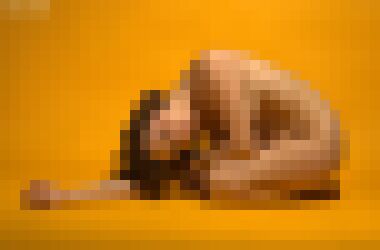 A nude young white woman with short brown hair kneeling with her body facing left, leaning forward until her right arm rests on the floor and draping her left over her knees, bowing and tilting her head as she looks at the camera with a neutral expression