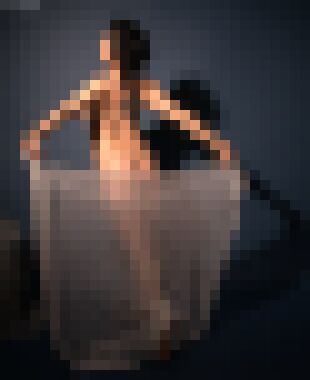 A back shot of a nude woman with curly black hair and a tattoo on her right shoulder blade arching her back and puching her left leg behind her, holding a sheer white shawl between her outstretched hands, glancing over her left shoulder with a neutral expression