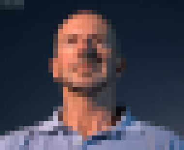 A bald white man with neat stubble looking above the camera with a neutral expression