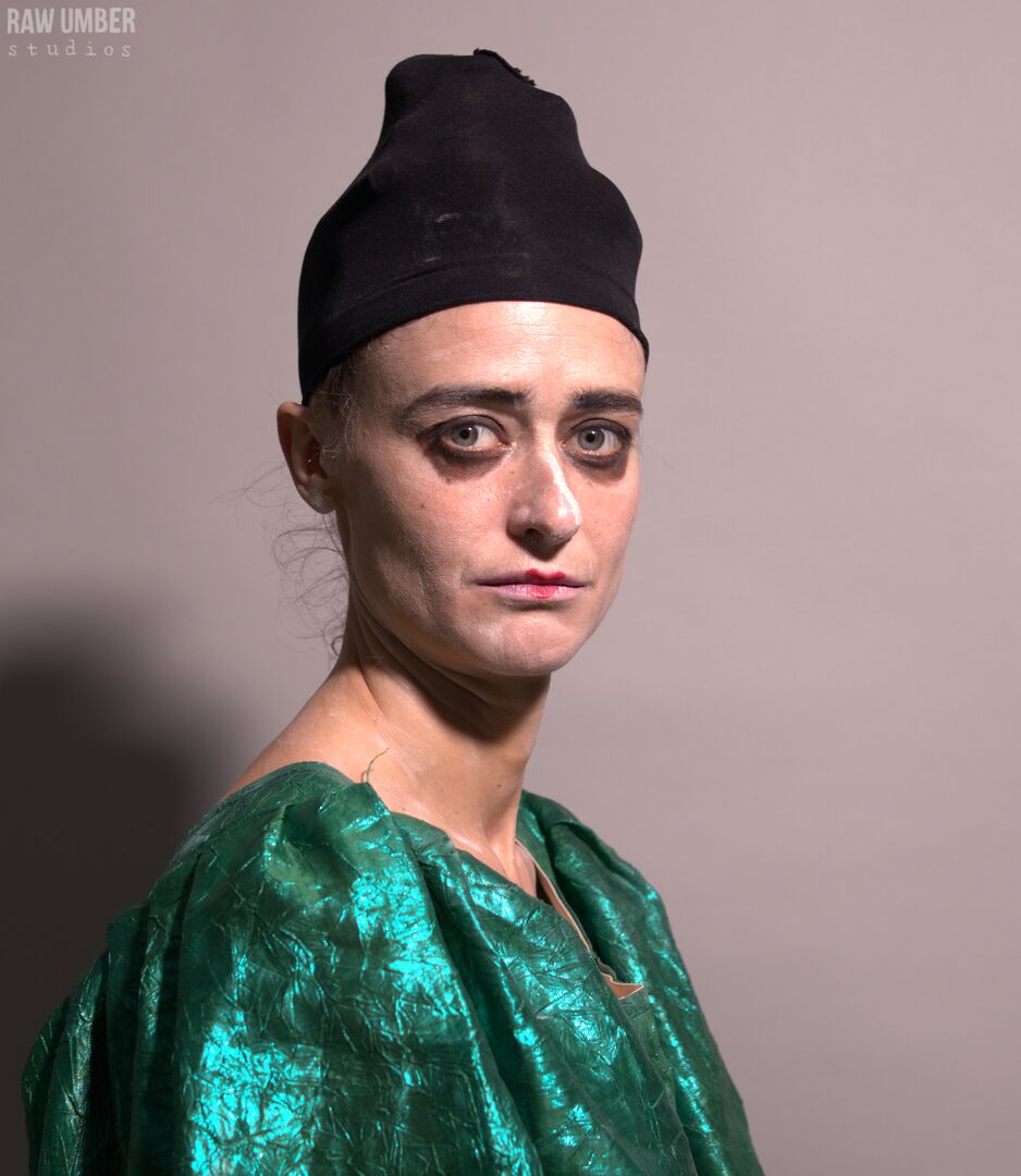 A woman with pale makeup, dark eyeliner, and a black head wrap looking over her right shoulder at the camera with a worried expression