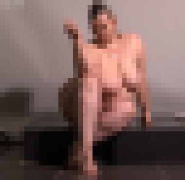 A nude white woman with a mohawk sitting on a black box with her hips cocked to the left, her left leg tucked under her right, her right arm resting on her knee palm upward, and her left arm supporting her, glancing at the camera with a slight smirk