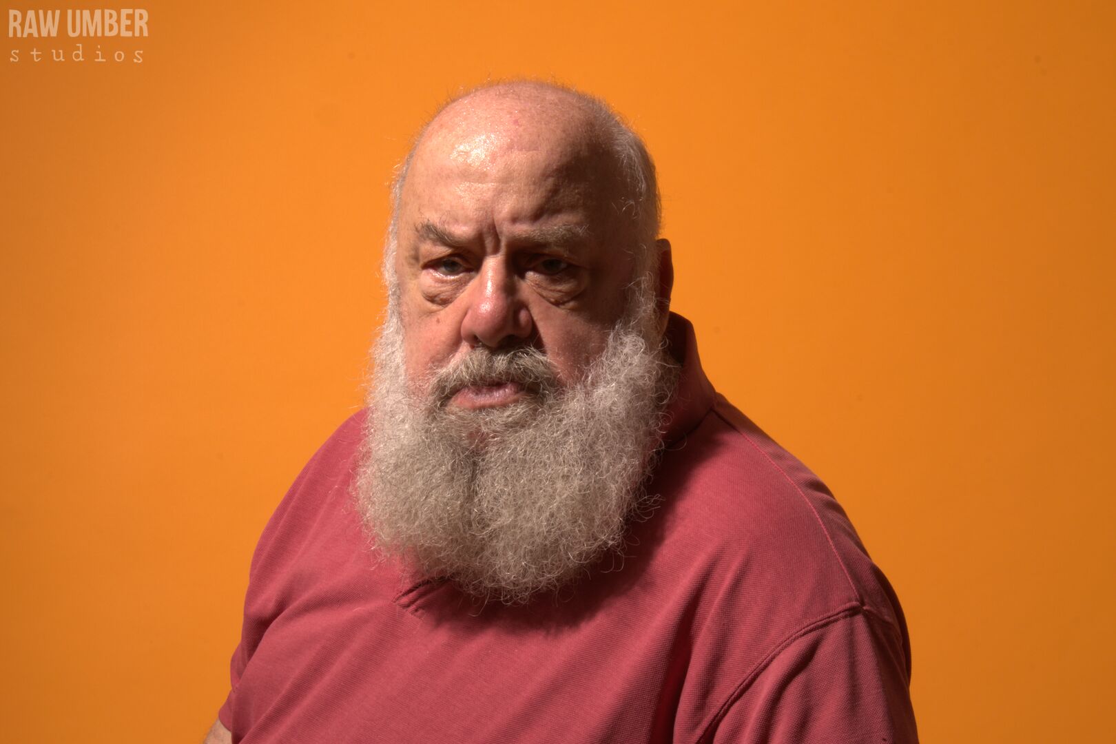 A bald white man with a bushy grey beard looking at the camera with a quizzical expression