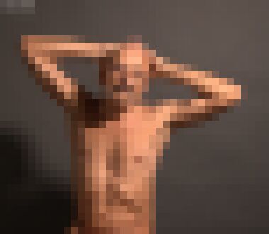 A nude, bald middle-aged man with glasses and a moustache holding his hands behind his head and looking straight at the camera with a neutral expression