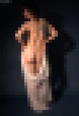A back shot of a nude woman with curly black hair and a tattoo on her right shoulder blade standing with a sheer white shawl draping over her calves, holding both her hands to her hips and glancing over her left shoulder with a neutral expression