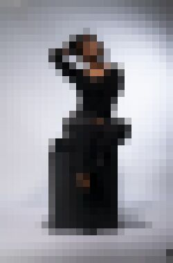 A black woman with thin dreadlocks sitting atop a black box, her left leg tucked under her right and her right hand behind her head, looking up and to the right with a neutral expression